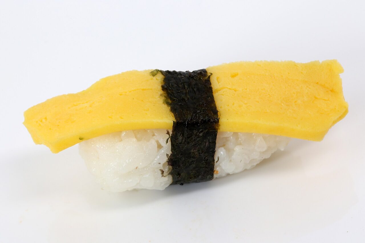 白い台の上に、卵焼きをのりの帯で巻いた握りずしがひとつ