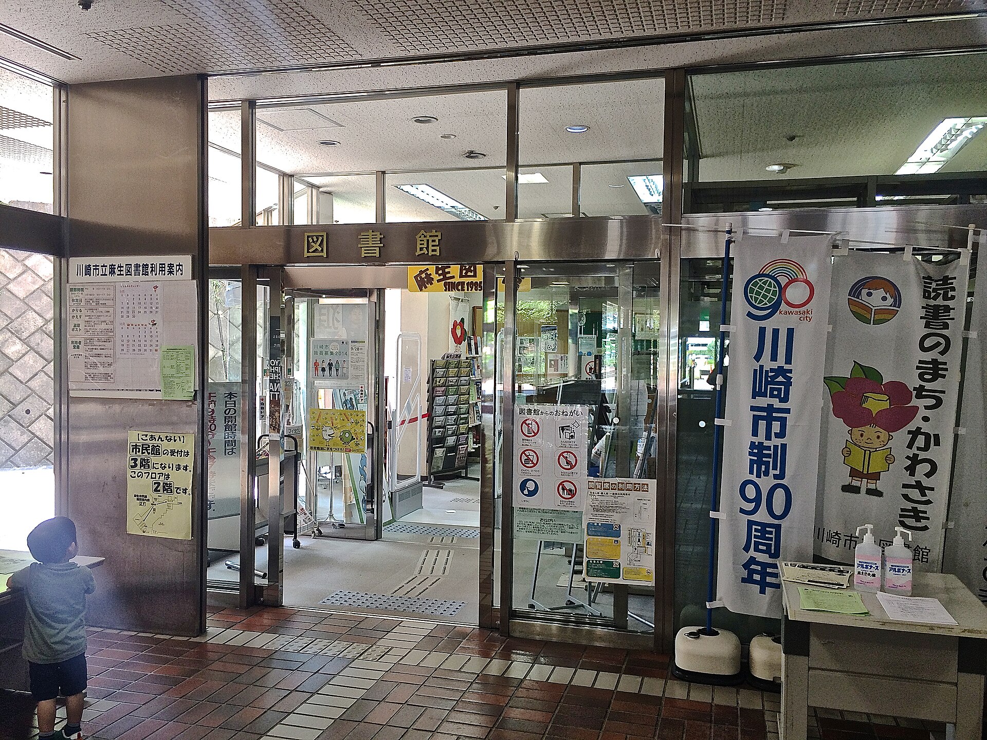 麻生図書館の入口。「図書館」の文字の上に、黄色い看板の「麻生図　SINCE 1985」が見える。左の柱には利用案内と「市民館の受付は3階。このフロアは2階です」の掲示