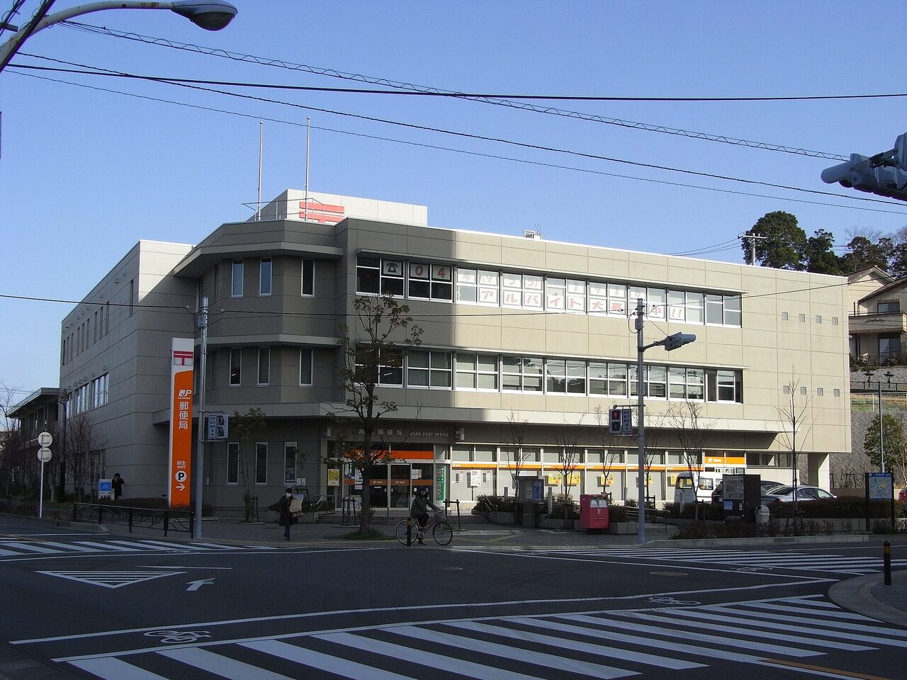 交差点の角に建つ、3階建ての灰色の麻生郵便局。入口の上に「麻生郵便局」の文字、左にオレンジ色の郵便局の看板が立つ