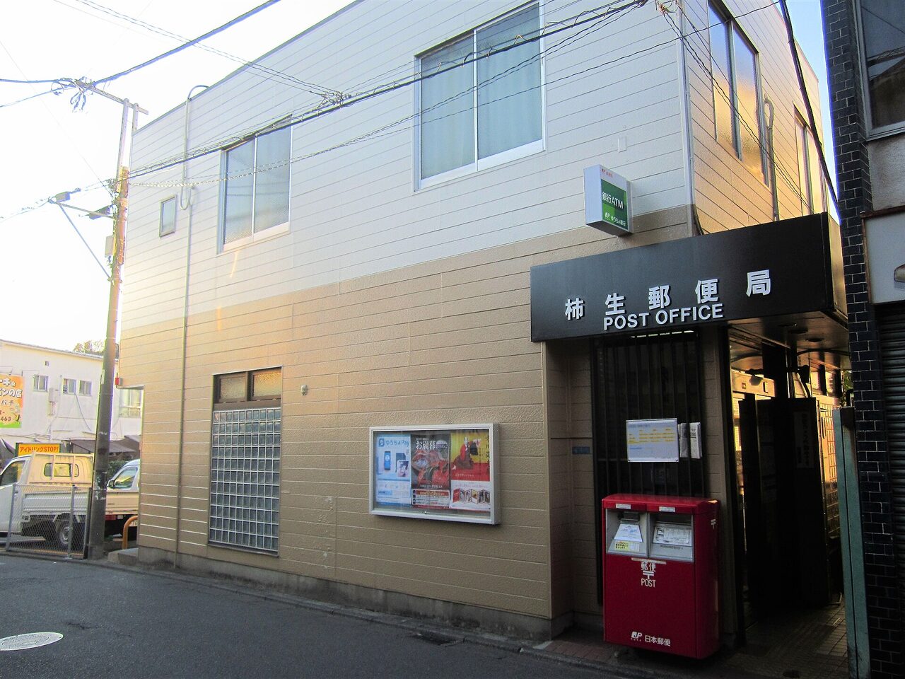 2階建ての建物の角に、黒い看板「柿生郵便局 POST OFFICE」。入口の前に赤い郵便ポストがある