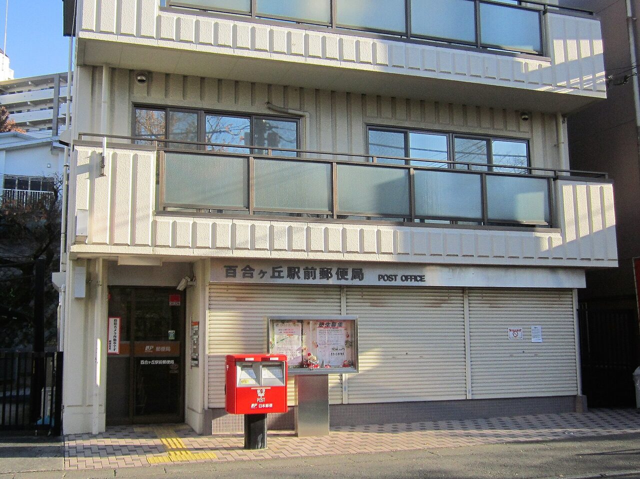 白い建物の1階に「百合ヶ丘駅前郵便局」の看板。シャッターが下り、手前に赤い郵便ポストが立つ