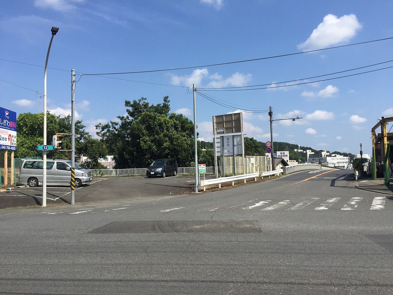 上麻生交差点。広い交差点の先に道路が続き、左に車の止まった駐車場、奥に緑の丘が見える