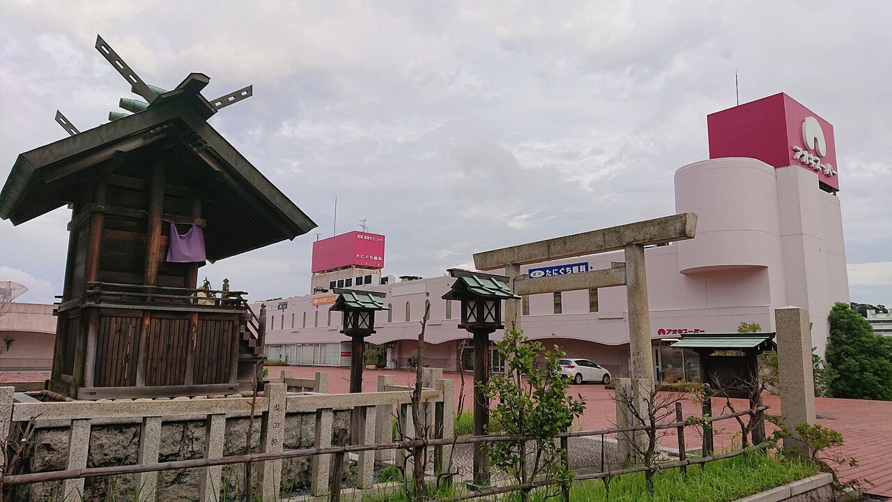 千代ヶ丘椿神社の小さな社と石の鳥居。奥に商業施設の建物が見える