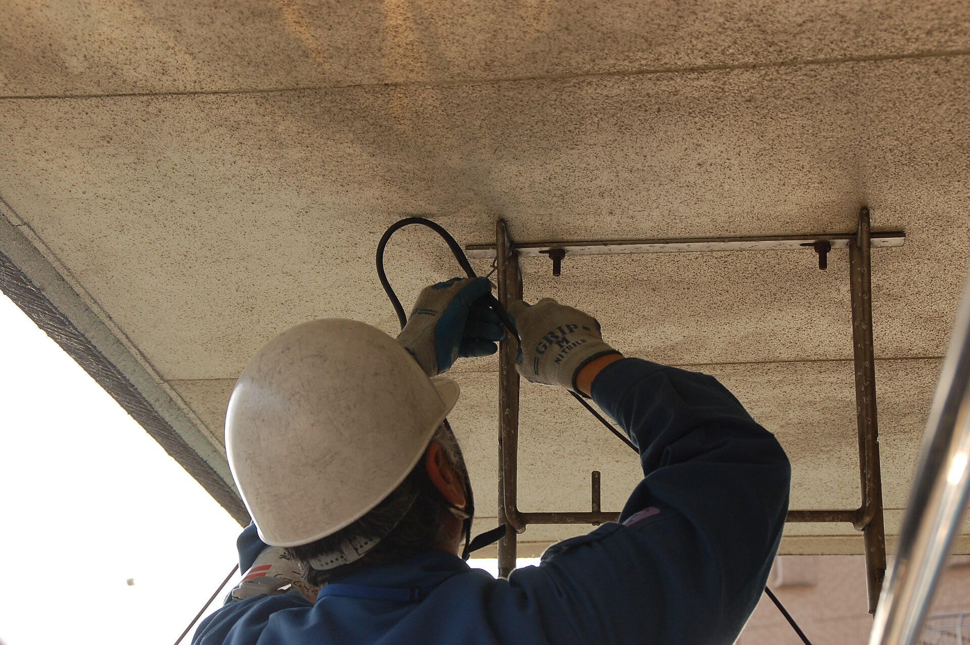 ヘルメットをかぶった作業員が、建物の軒下で、黒いケーブルを金具に通している後ろ姿
