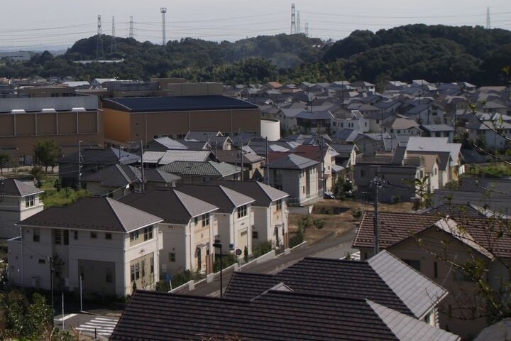 高台から見たはるひ野の住宅地。灰色や茶色の屋根の家が並び、奥に大きな建物と緑の丘、送電線の鉄塔が見える