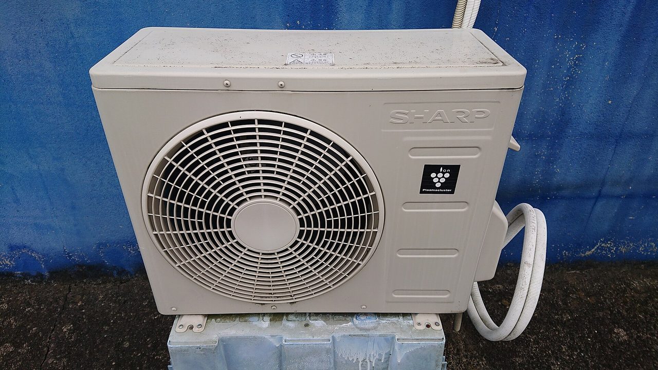 青い壁の前の台に置かれた、ファンの丸い格子がある室外機。横から白い配管がのびている