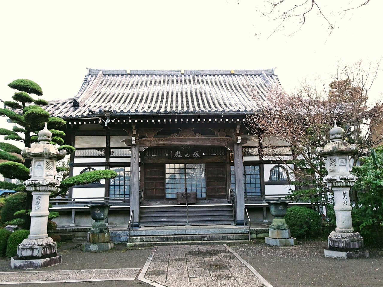 瓦屋根の寺の本堂。正面に石段と扁額があり、左右に石灯籠、左に松の木が立つ（2024年11月撮影）