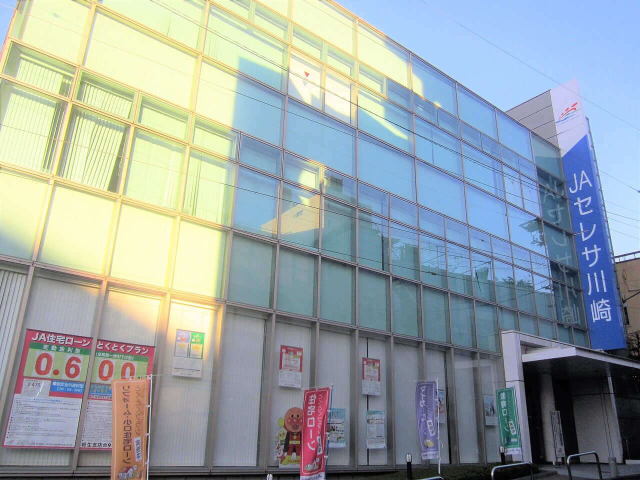 ガラス張りの建物の右に、縦長の青い看板「JAセレサ川崎」。入口の前にのぼりが並ぶ