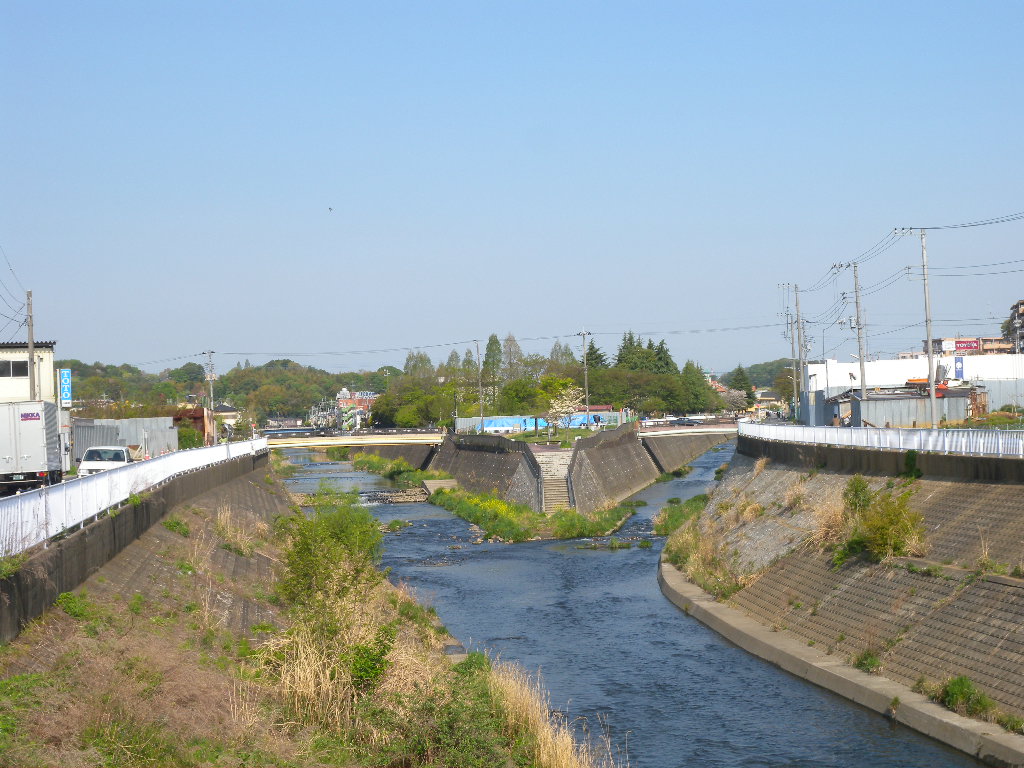 コンクリートの護岸にはさまれた川。奥で2つの流れが合わさり、遠くに低い山並みと家並みが見える