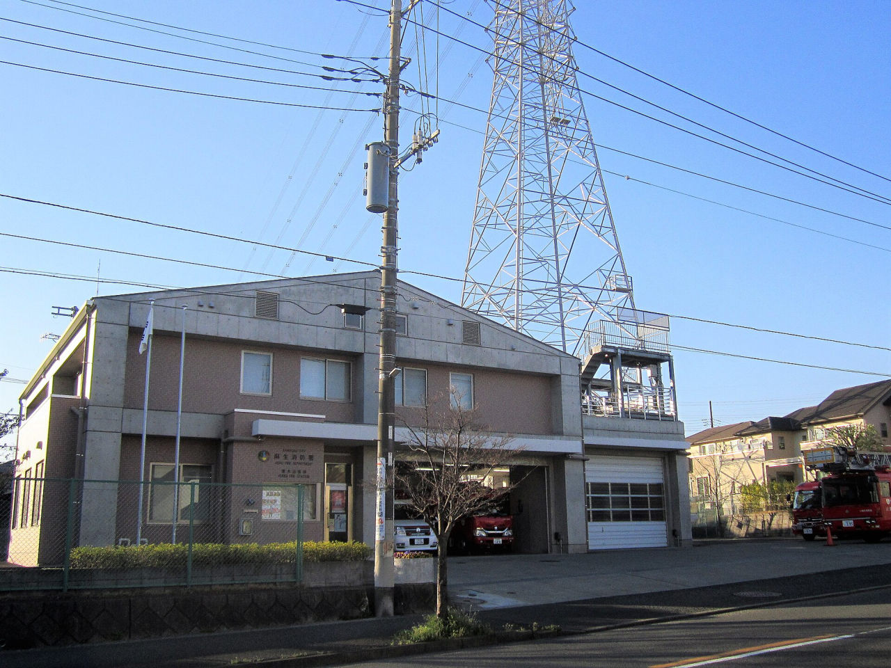 栗木台にある麻生消防署栗木出張所。2階建ての建物の車庫に消防車が止まり、うしろに送電線の鉄塔が立っている