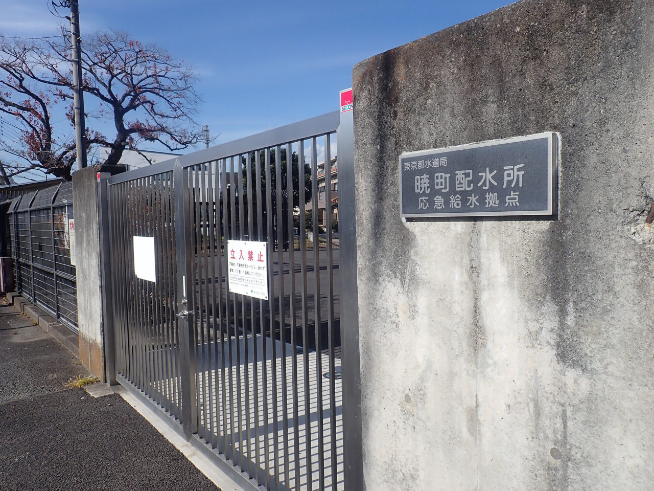 コンクリートの門柱に「東京都水道局 暁町配水所 応急給水拠点」と書かれた看板。横に閉じた金属の門がある