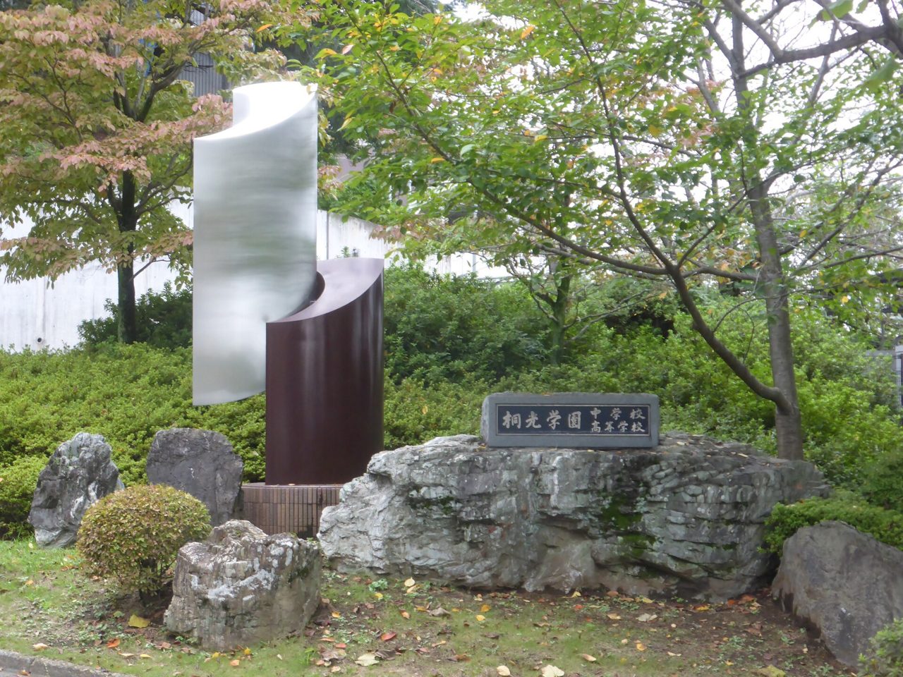 桐光学園中学校・高等学校の入口。石の台に校名の札があり、うしろに金属の像と木々が立っている
