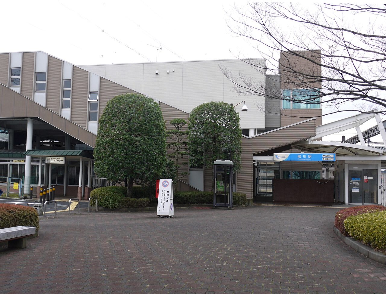 駅舎の前に、れんが色の舗装の広場と植え込みがある。奥の入口の上に駅名の看板が見える