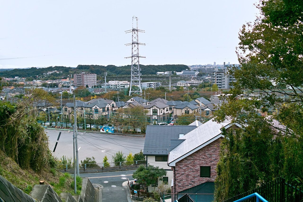 坂の上から見下ろした住宅地。手前に下り坂の道、中ほどに広い駐車場、奥に鉄塔と丘が見える