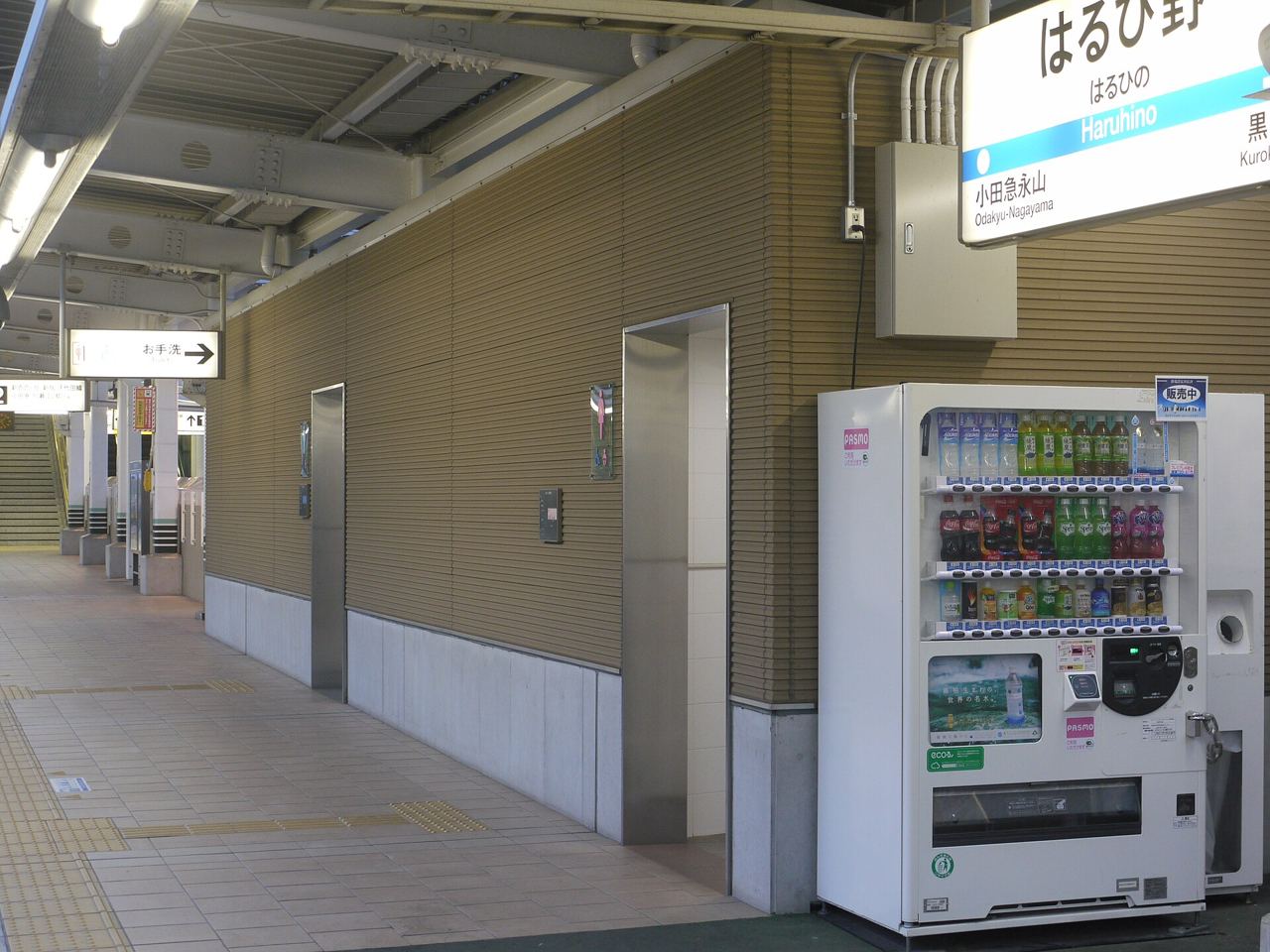 はるひ野駅のホーム。壁にトイレの入口が並び、手前に飲み物の自動販売機と、駅名の看板がある