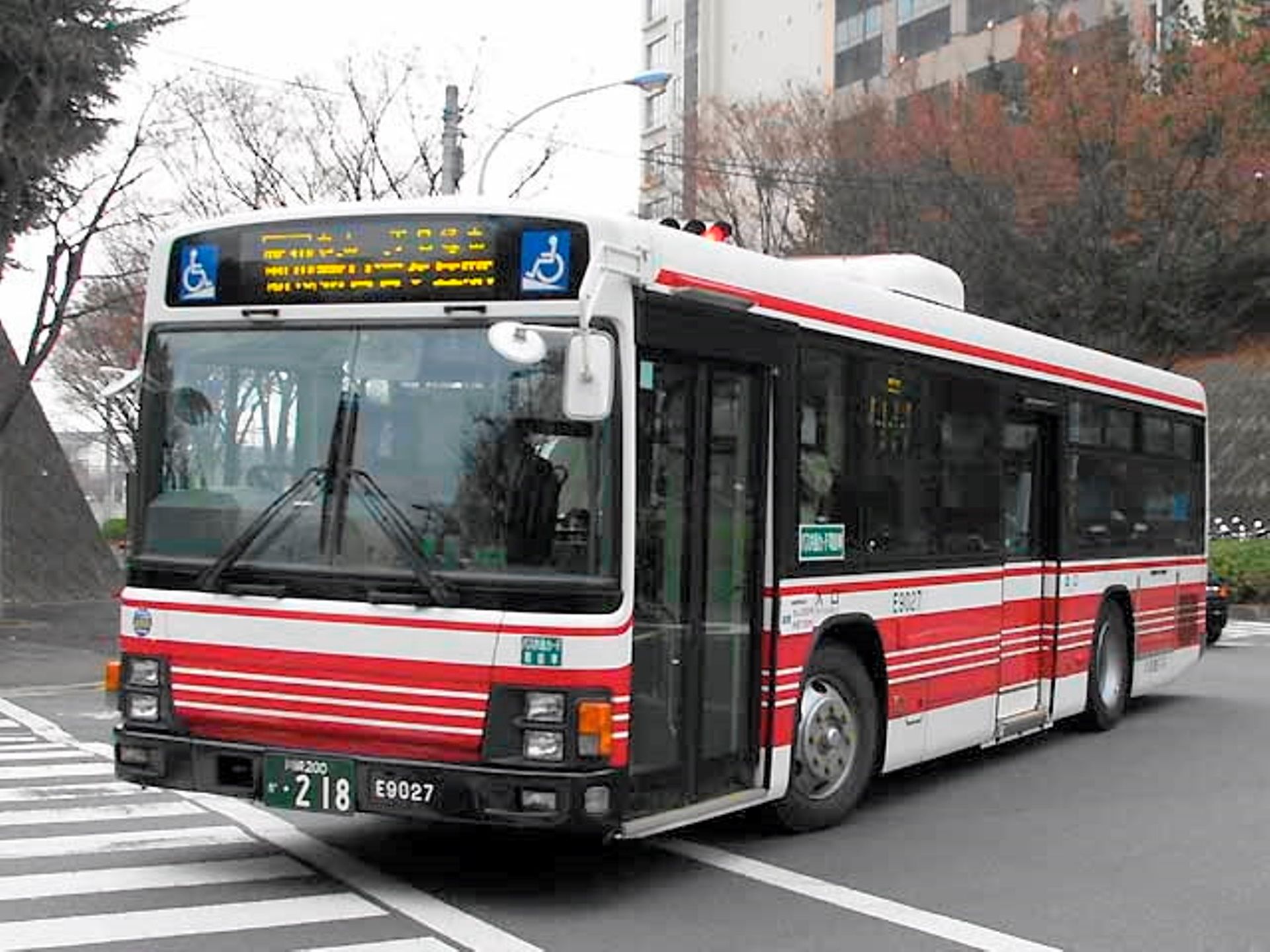 赤と白の路線バスが、横断歩道の手前を走っている。後ろに紅葉した木と建物