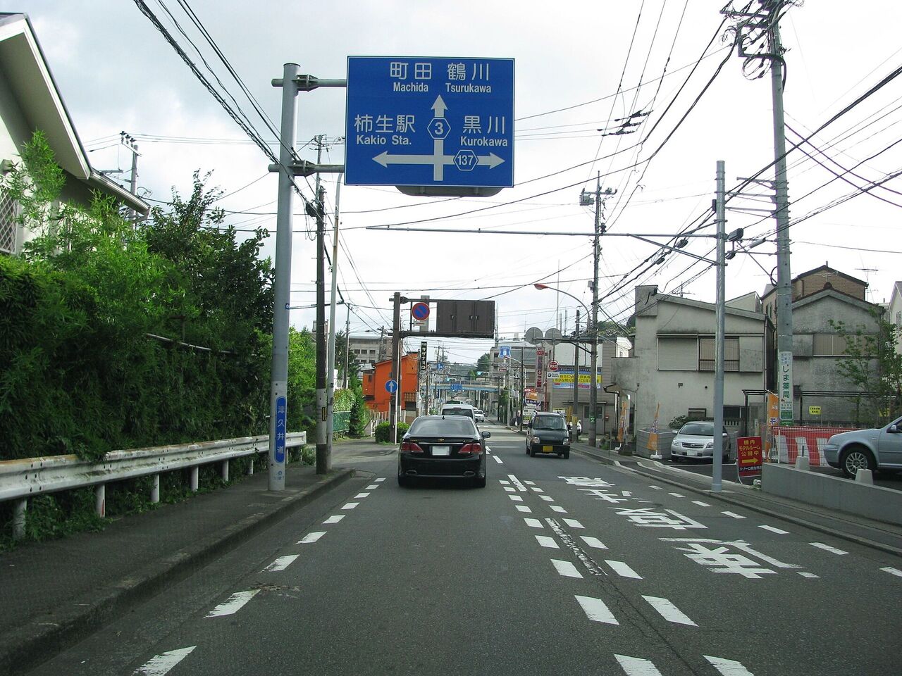 片平の県道。道路の上に「町田・鶴川」「柿生駅」「黒川」と書かれた青い案内標識があり、道の両側に住宅が並ぶ