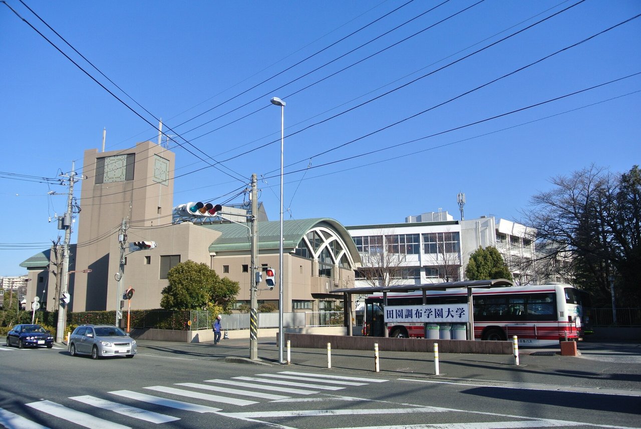 交差点の角に、大学の建物と、アーチ形の屋根の建物。歩道の横に「田園調布学園大学」の看板を付けたバス停の屋根があり、奥にバスが停まっている