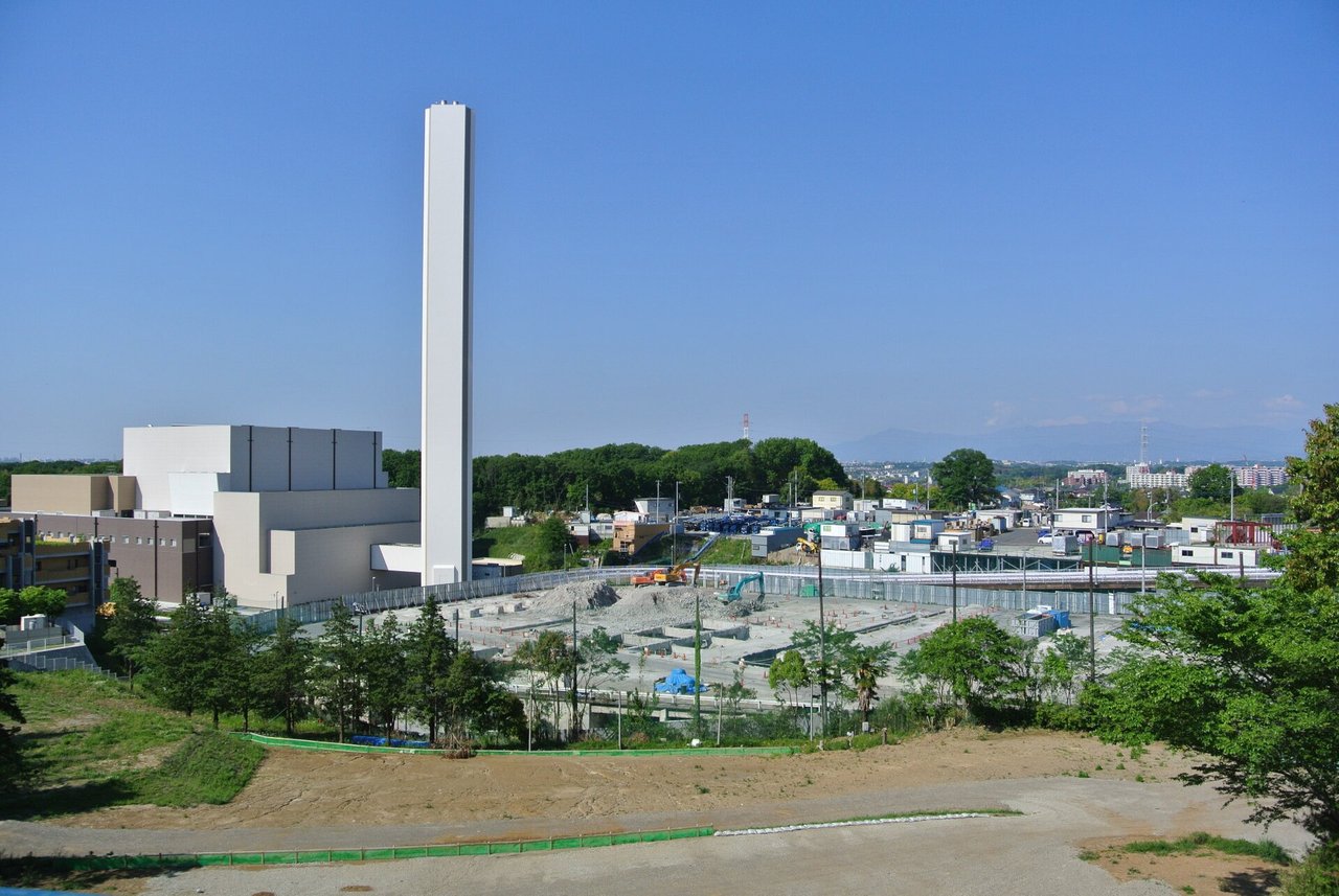 丘の上から見た、高い白い煙突と四角い建物。手前は整地中の広い土地で、奥に住宅と山並みが見える