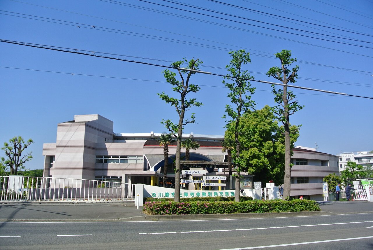道路の向こうに、薄い茶色の3階建てほどの建物と、施設の名前が書かれた低い塀。手前に街路樹が並ぶ（2013年撮影）