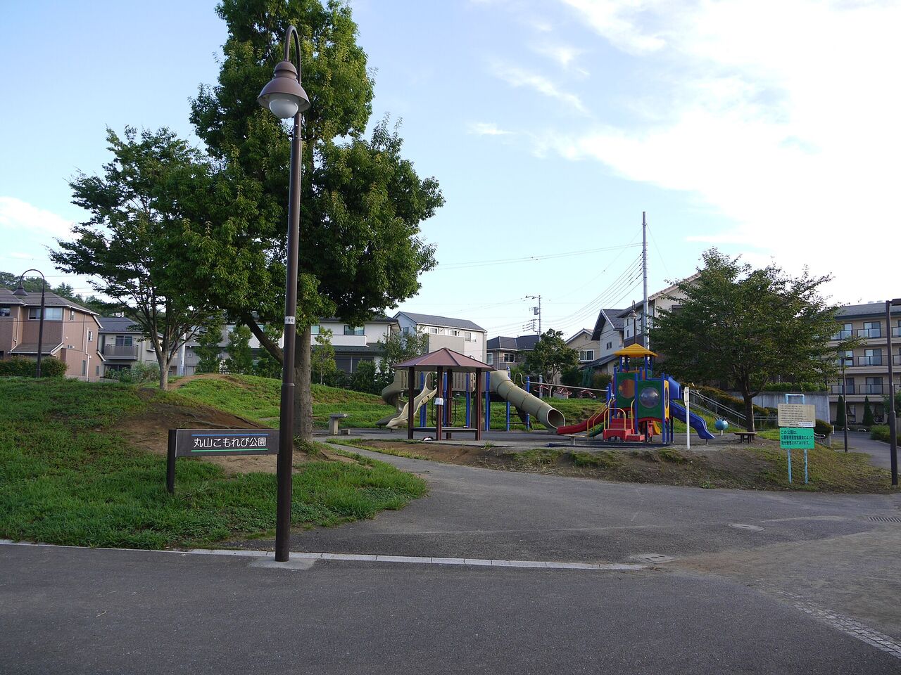 芝生の小山と遊具のある公園。まわりに2階建ての家と集合住宅が並んでいる