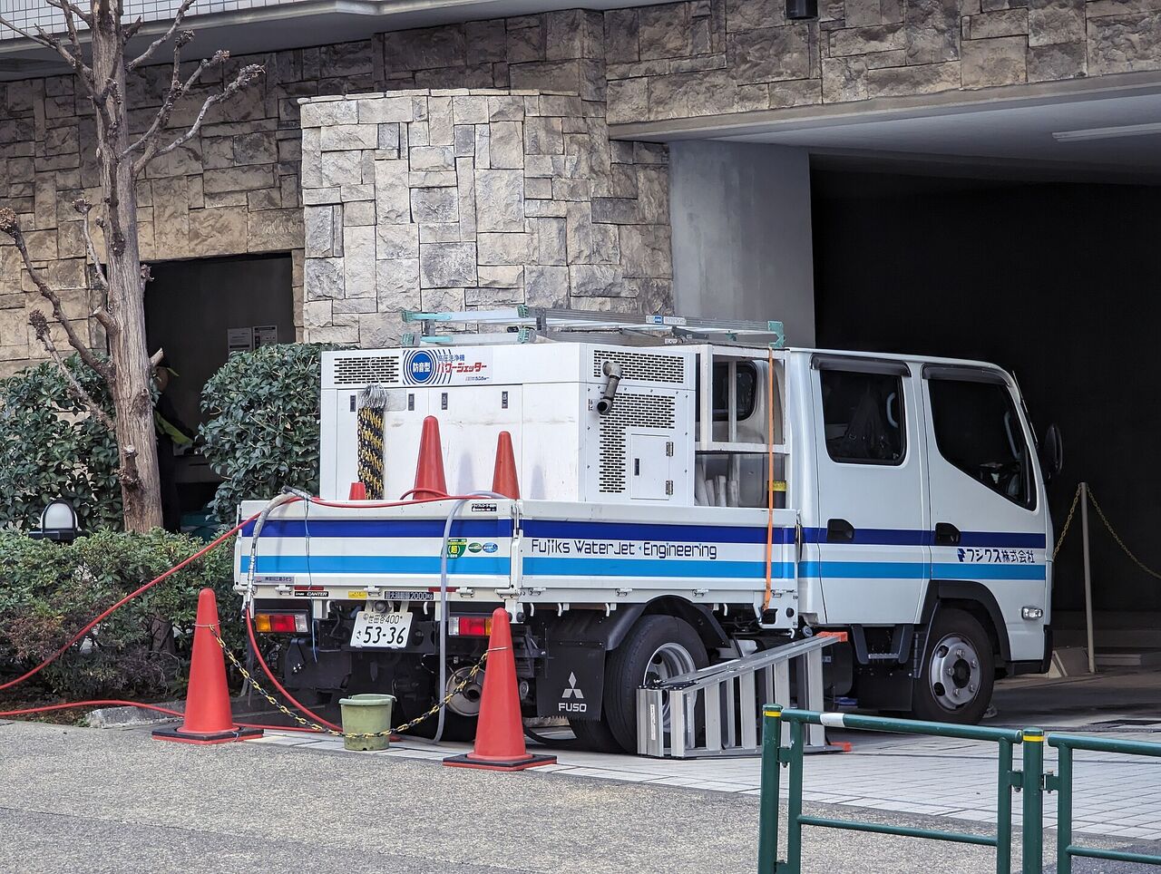 ビルの前に止まった、高圧洗浄用の機械をのせた作業車。まわりに赤い三角コーンが置かれている