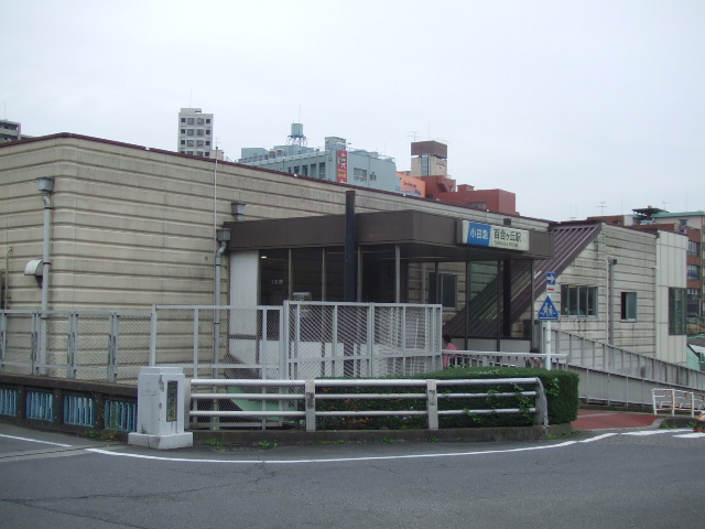 ベージュの壁の駅舎の入口。屋根の上に小田急の青い看板と「百合ヶ丘駅」の文字があり、手前に柵と植え込みがある