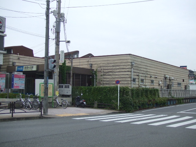 平屋の駅舎の左端に、小田急の青い看板と「百合ヶ丘駅」の文字。手前の歩道に自転車が止まり、横断歩道がある