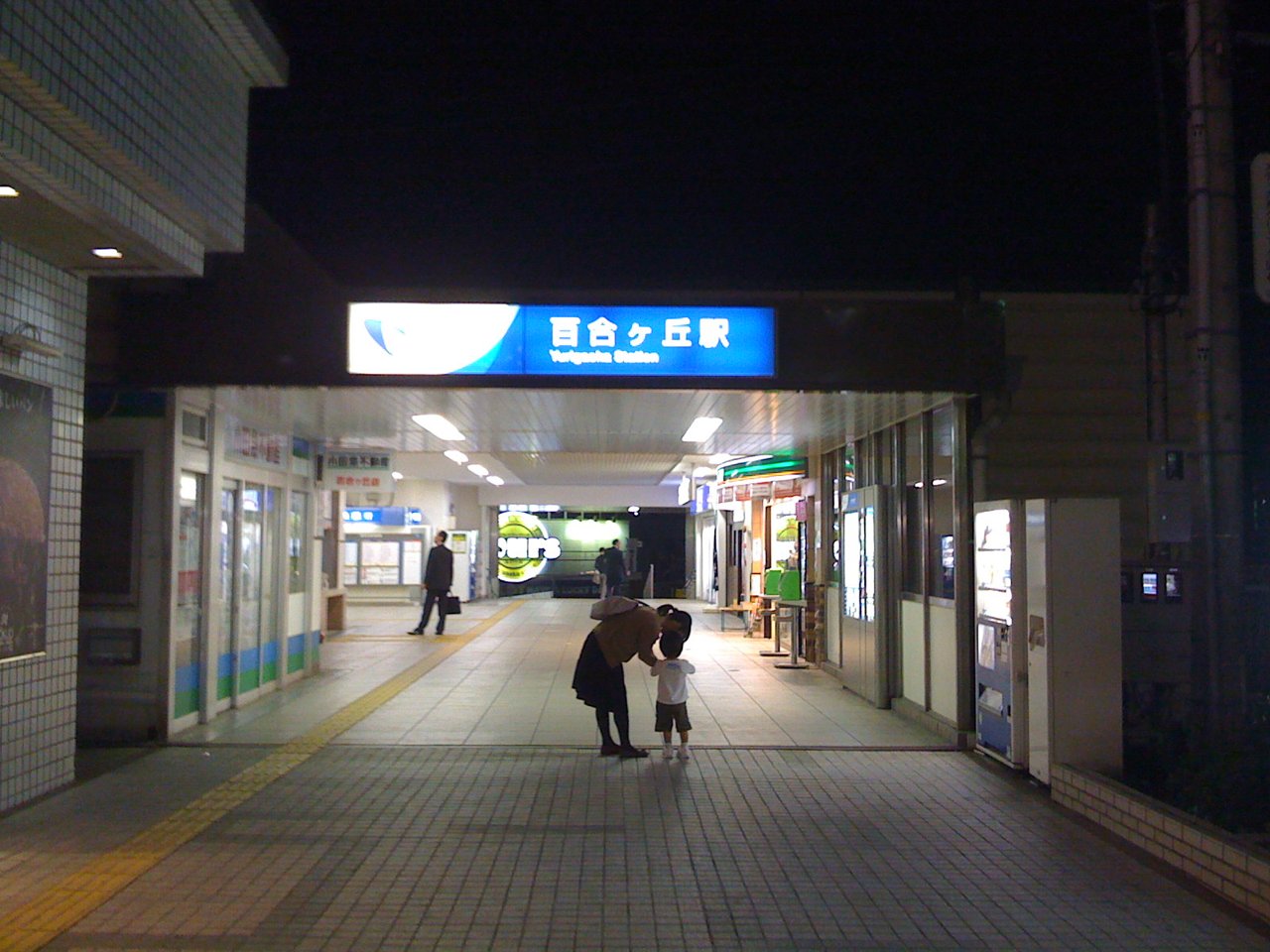 夜の百合ヶ丘駅の入口。青い「百合ヶ丘駅」の看板の下に明るい通路がのび、右側に店の明かりと自動販売機が並ぶ(2008年撮影)
